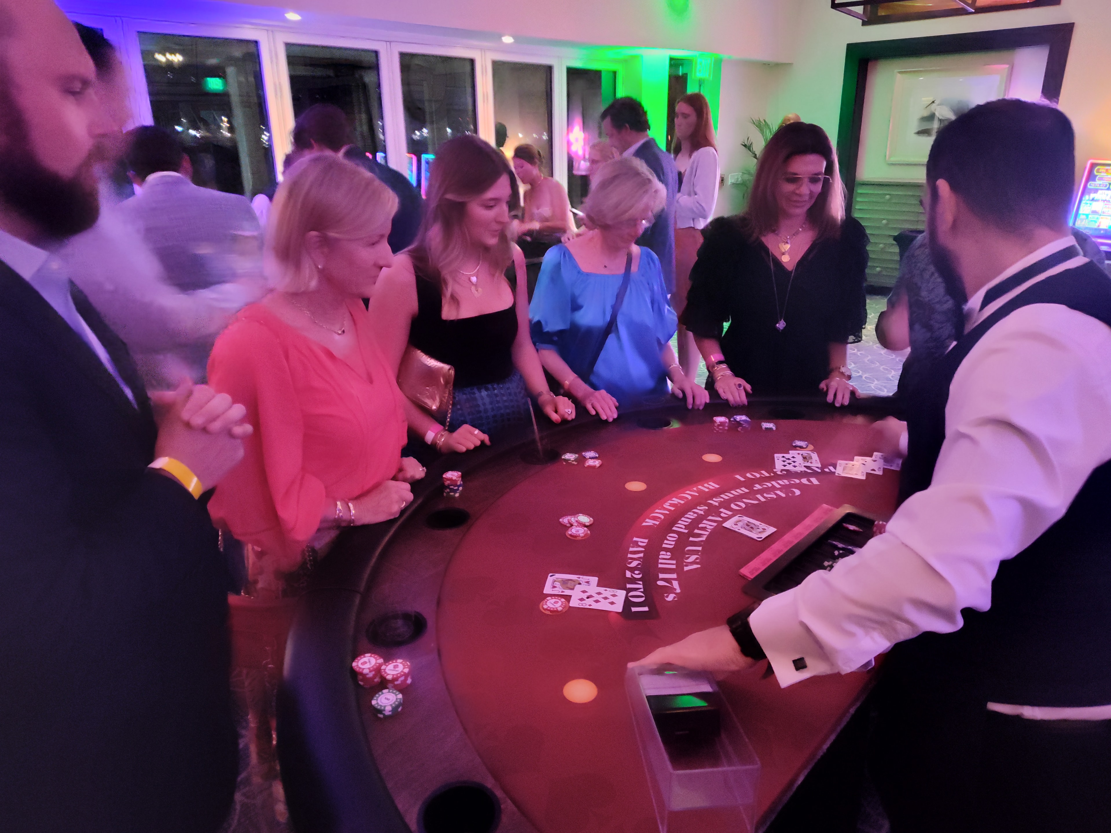 Guests in formal attire playing blackjack at a Casino Party USA table with green uplighting in Miami