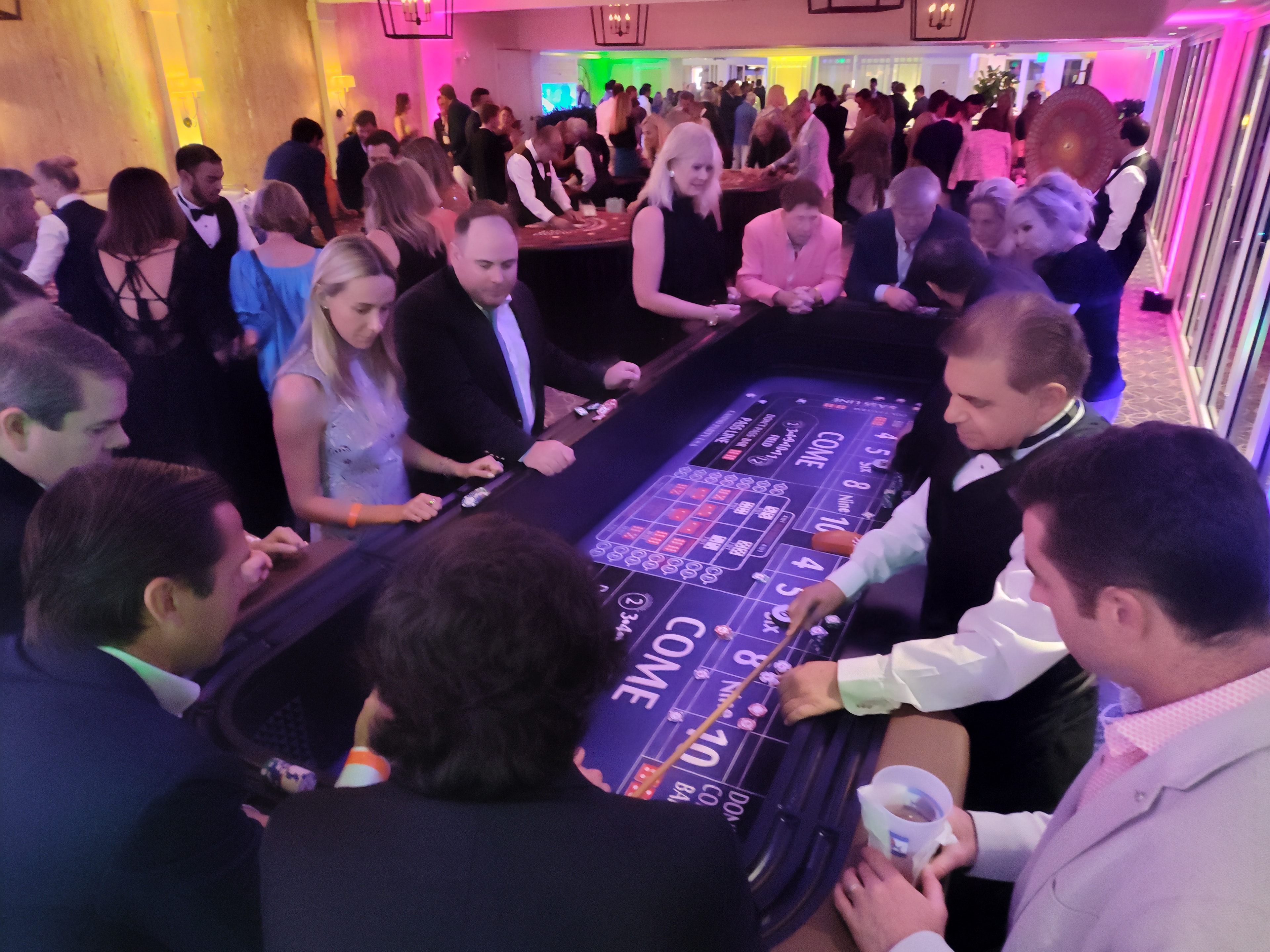 Large crowd of guests around a Casino Party USA craps table with colorful uplighting in Miami
